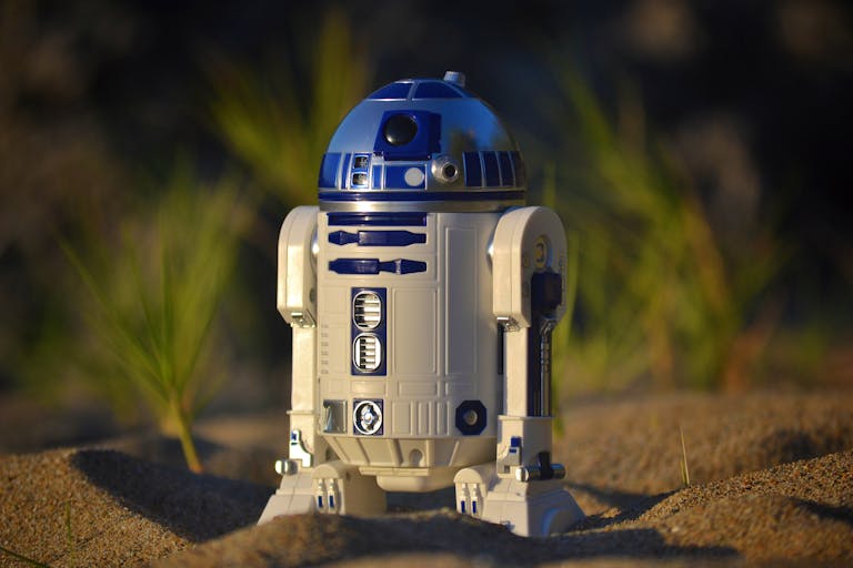Close-up of an R2-D2 toy standing on sand, set against a green background in bright daylight.